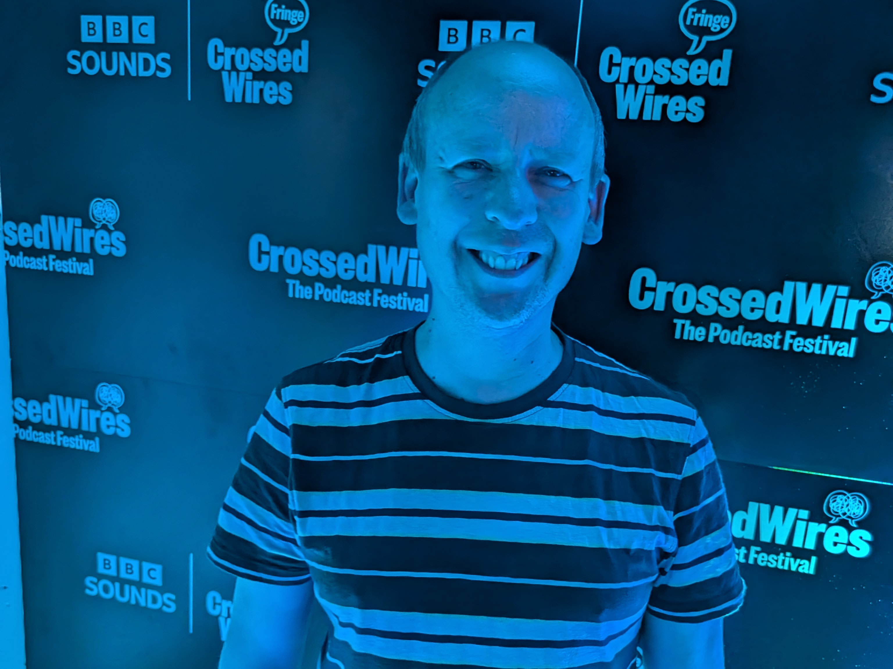 Close up of Andy in a blue lit room, in front of a wall featuring the BBC Sounds and Crossed Wires Fringe logos